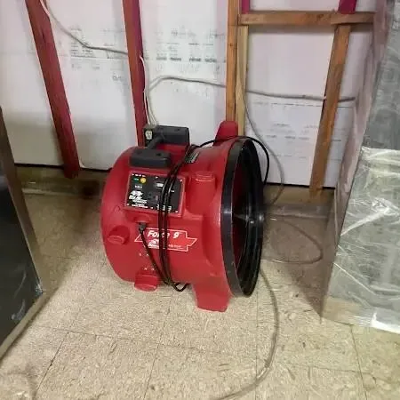 A red air mover drying a room with the lower wall cut open and plastic sheeting hung after a floor-level leak