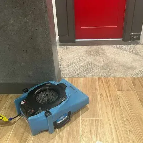 An air mover blowing across a wood-look tile floor beside an open doorway during drying